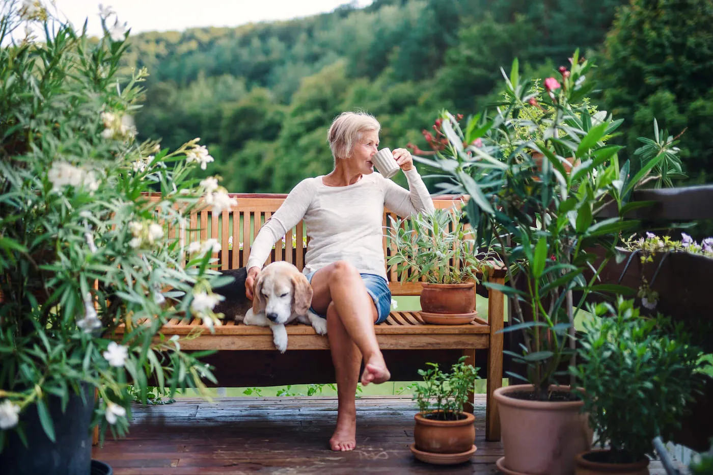 An elderly woman drinks from a cup while sitting on a bench next to her dog with trees in the background.