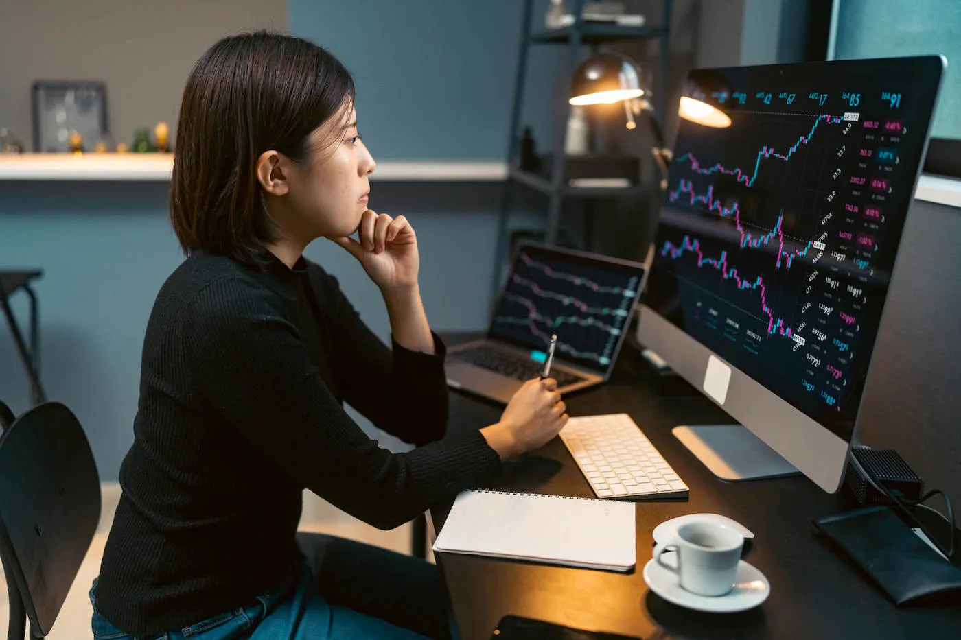 A woman wearing a black shirt holds a pen while looking at a stock chart on her computer screen with her laptop next to it.