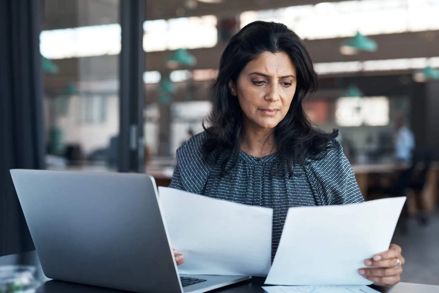 A woman wearing a black shirt holds two documents in her hand while across from her laptop.