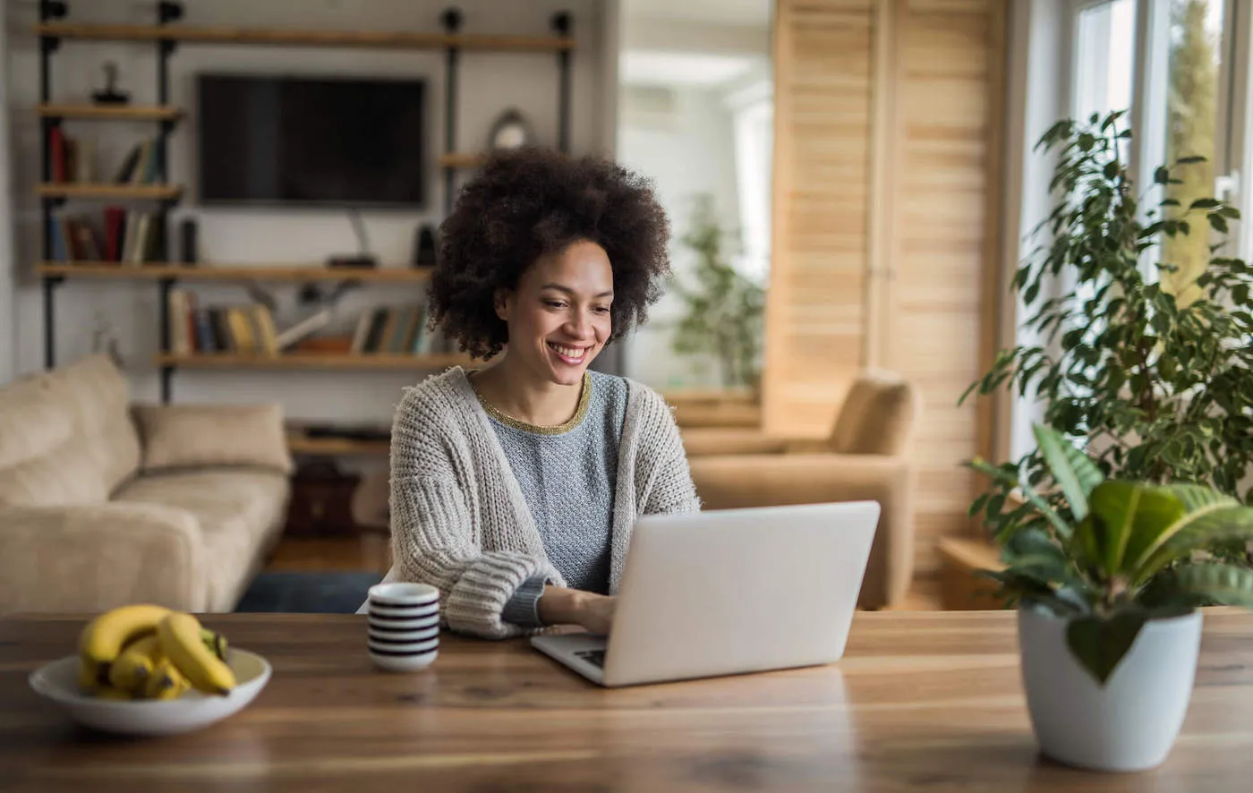 A woman wearing a gray sweater smiles while using her laptop at home.