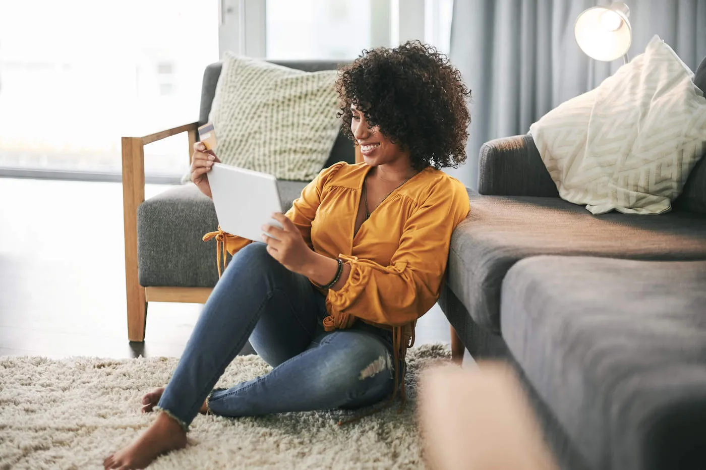 A woman wearing a yellow shirt smiles at her tablet while holding her credit card.