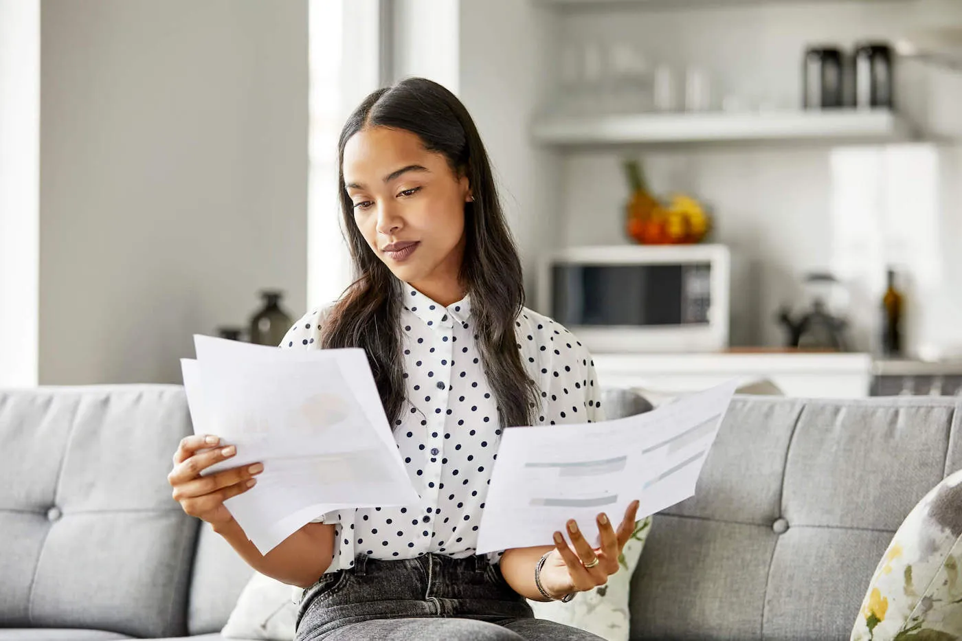 A woman is sitting on the couch wearing a polkadot shirt while looking at documents.