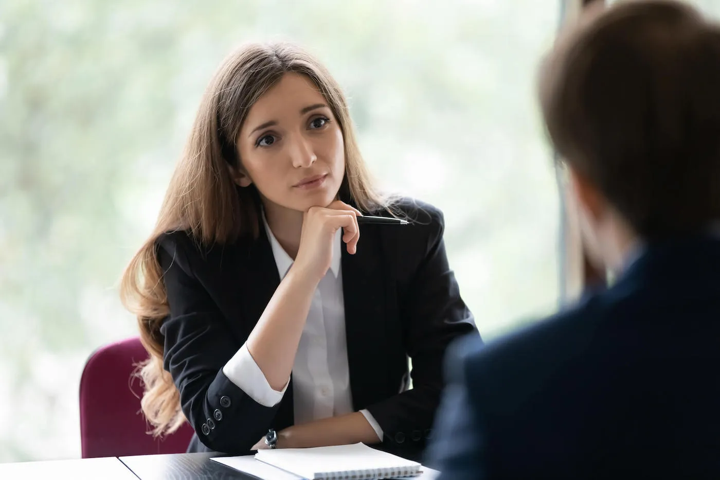 A woman wearing a suit listens to a man from across the table while holding a pen in her hand.