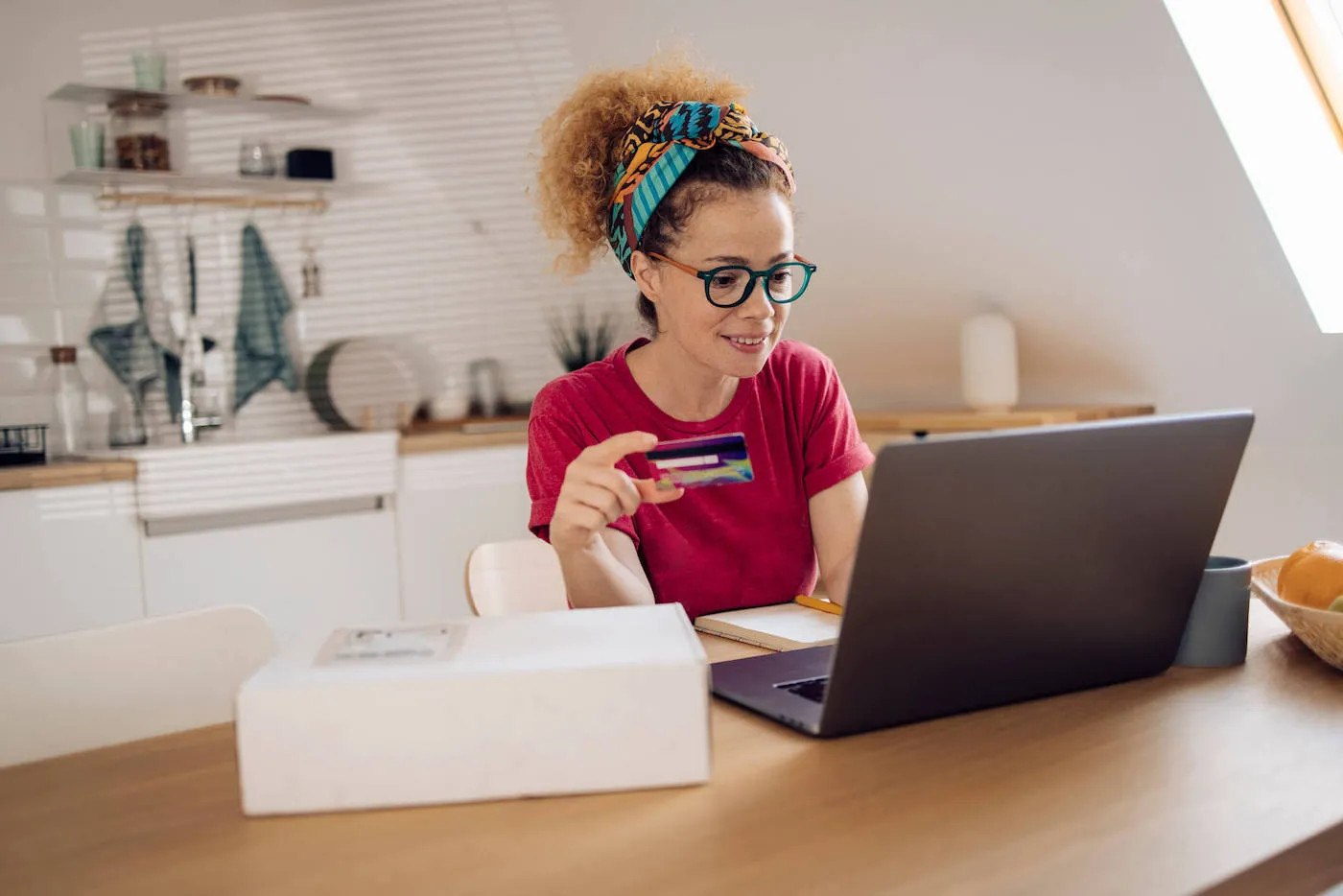 A woman wearing a red shirt and glasses uses her laptop computer while holding her credit card.