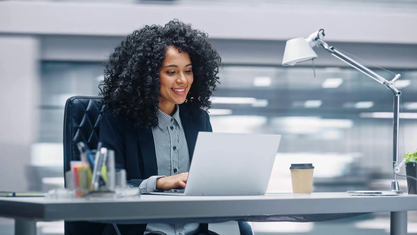 A woman wearing a suit smiles at her laptop computer screen while sitting down at her office desk.