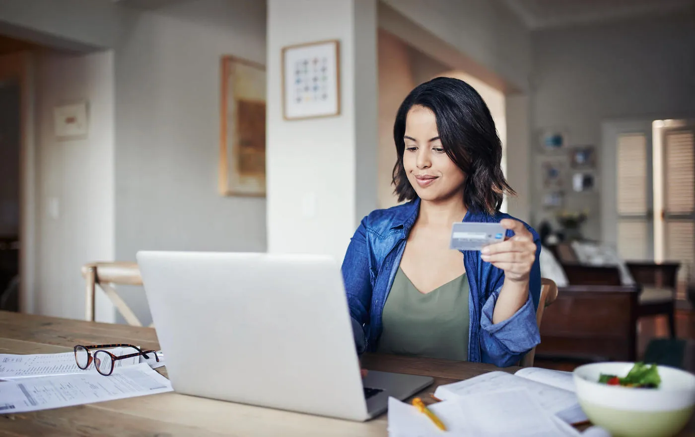 A women is typing on her laptop that is surrounded by documents as she holds her credit card in her had at the kitchen table.