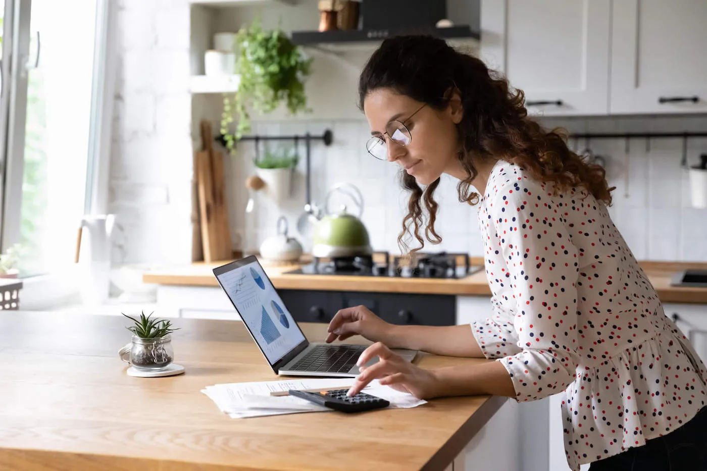 A women wearing a white polka dot shirt is using her calculator and laptop at the kitchen table.