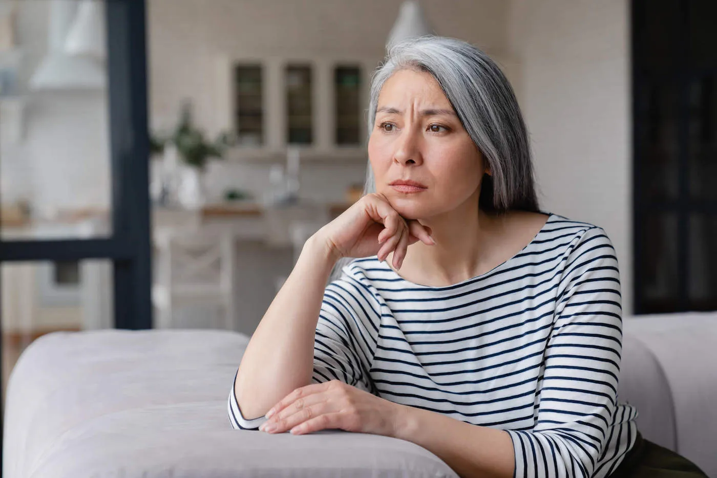 A women wearing a striped shirt is sitting on the couch leaning on her hand as she looks outside her window.