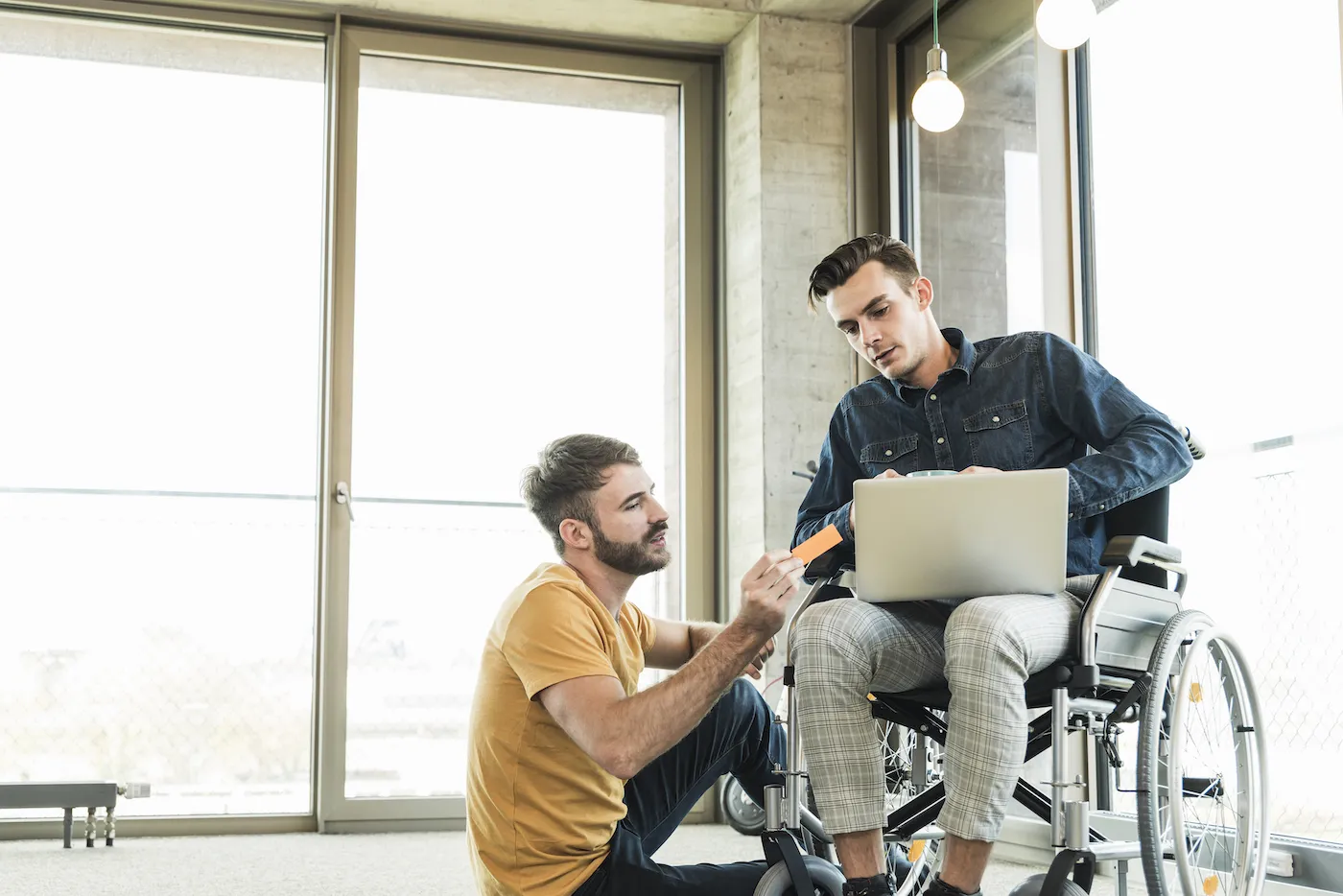 Young businessman in wheelchair and colleague using laptop in office.