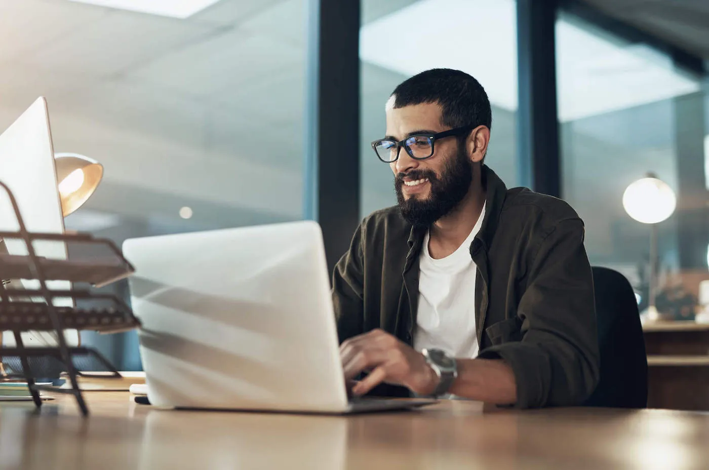 A young man wearing glasses and a green shirt is smiling as he types on his laptop at the office.