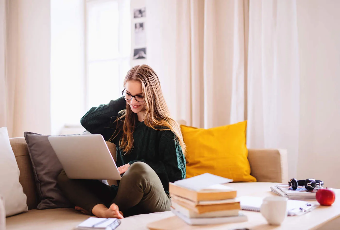 A young blonde student wearing glasses is sitting at the couch while smiling at her laptop with her books on a table beside her.