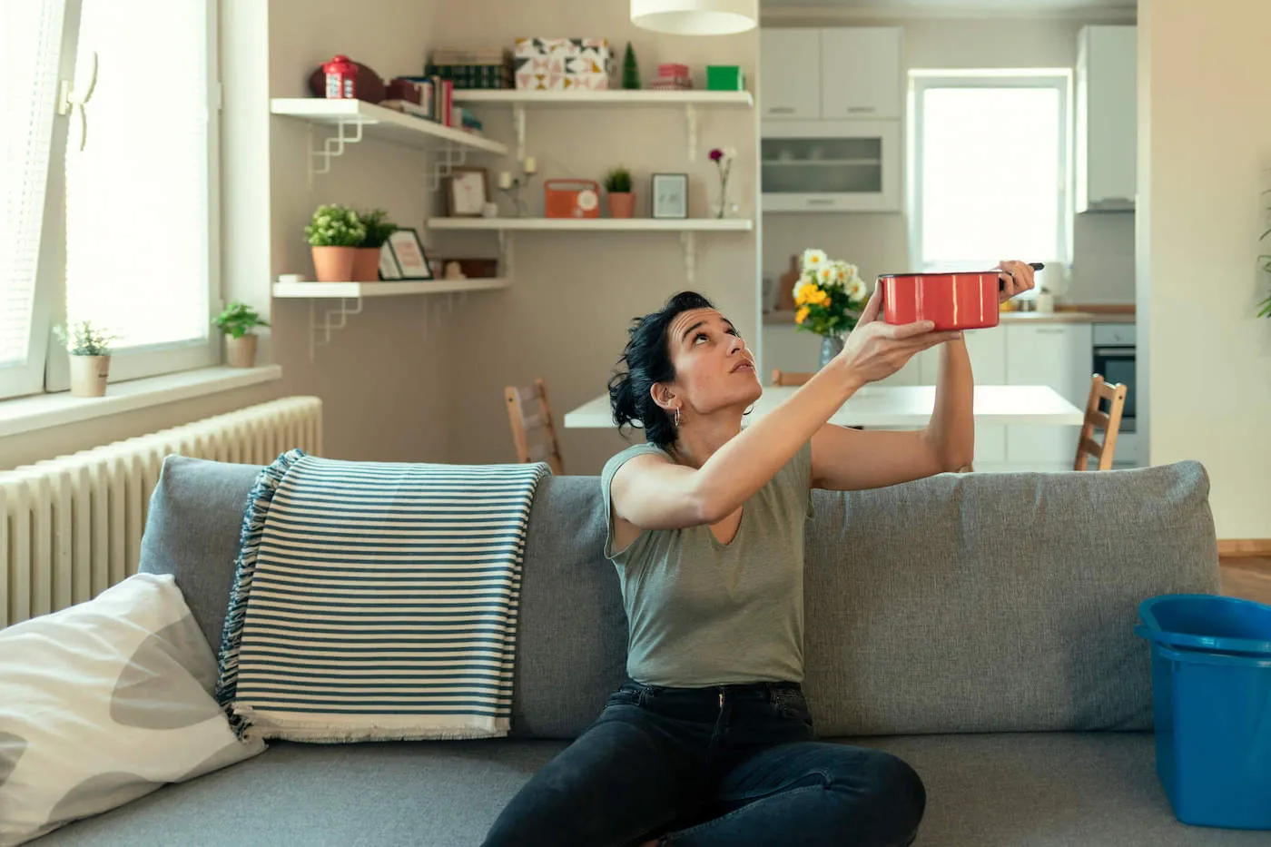 A young woman sitting on the couch holds up a pan to store the water dripping from the roof of her home.