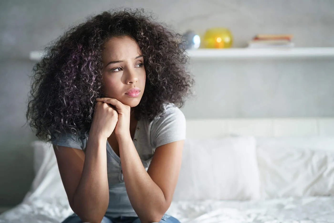 A young woman sits up and looks out the window of her bedroom looking sad.