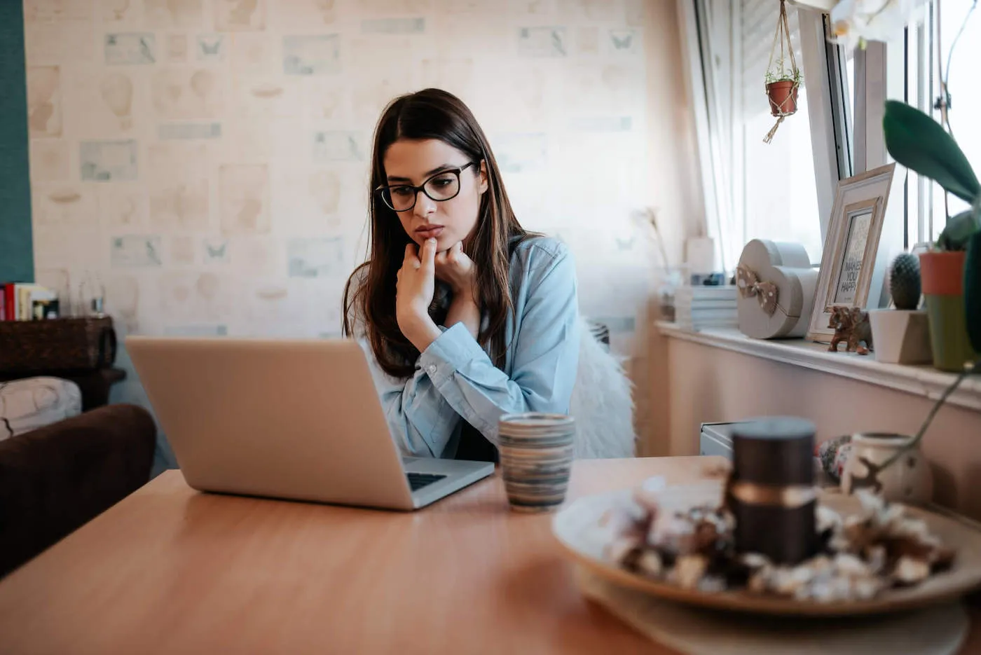 A young woman wearing glasses looks at her laptop screen while her hands are on her chin.