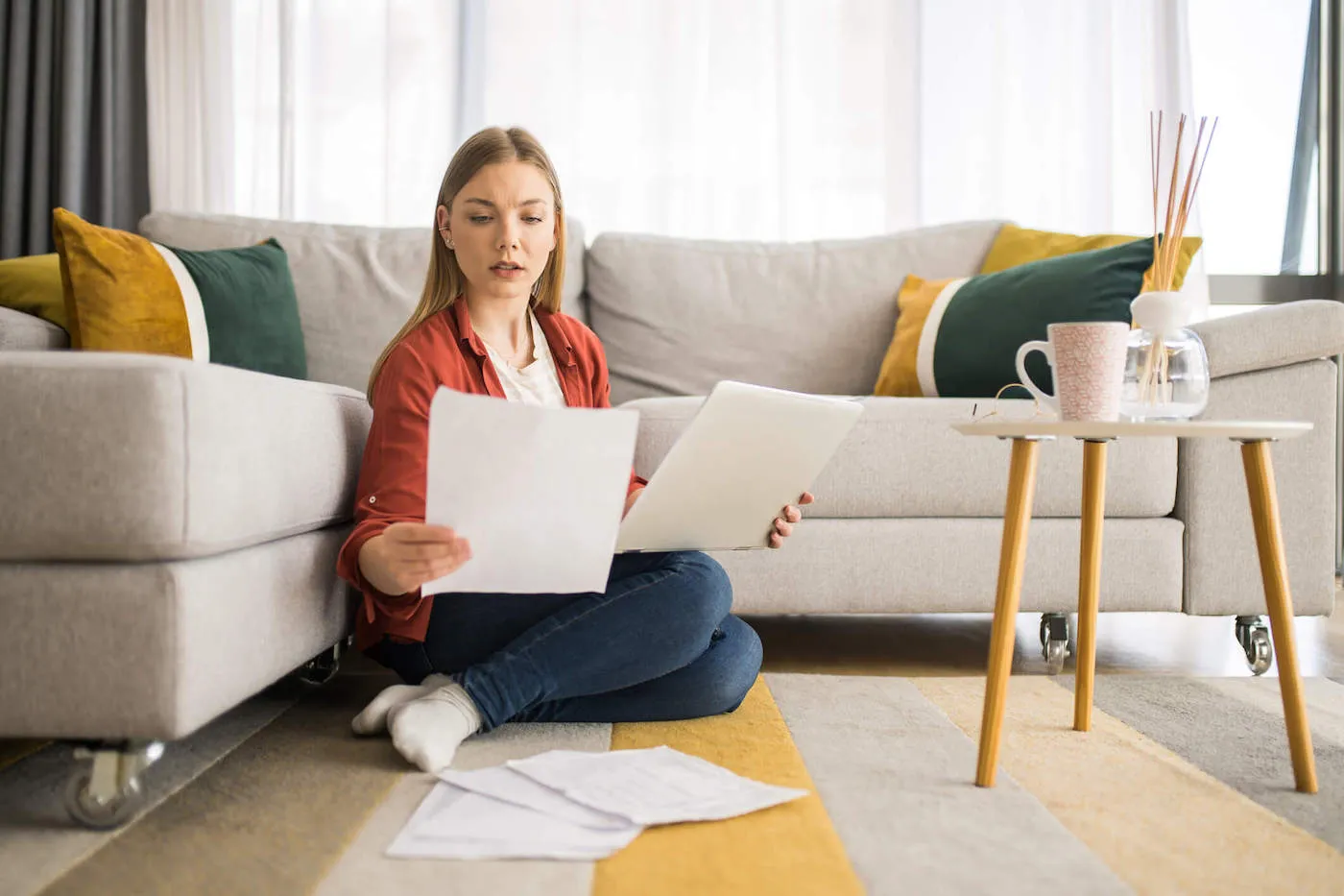 A young woman sits on her living room floor while looking at documents.