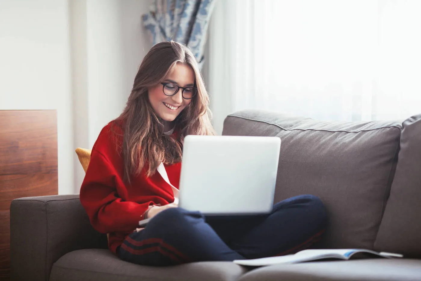 A young woman smiles at her laptop while sitting on the couch.