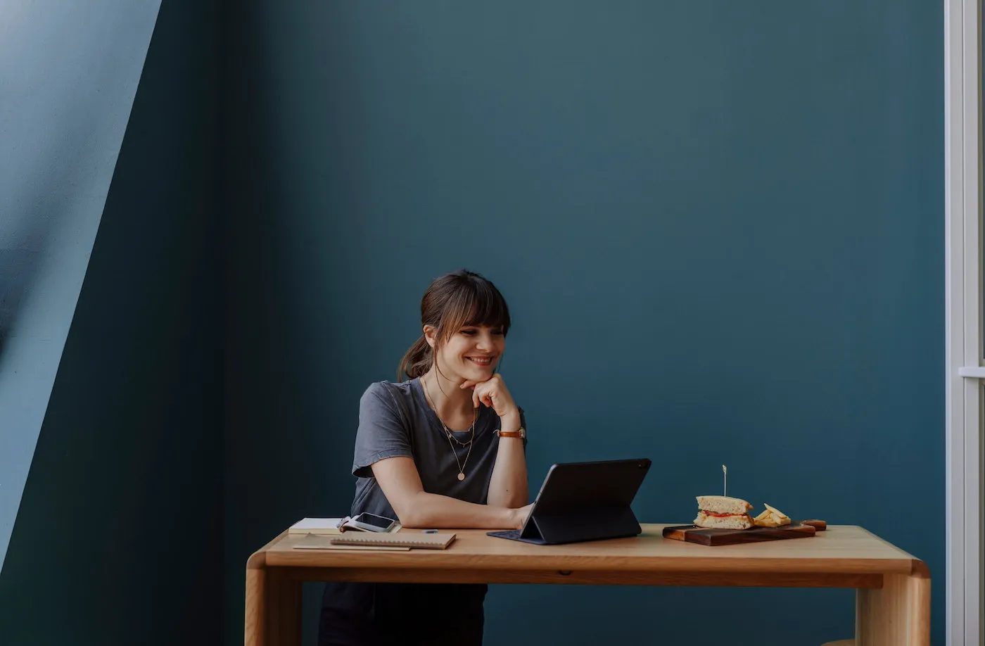 A young woman smiles while using her tablet with her sandwich right next to it.