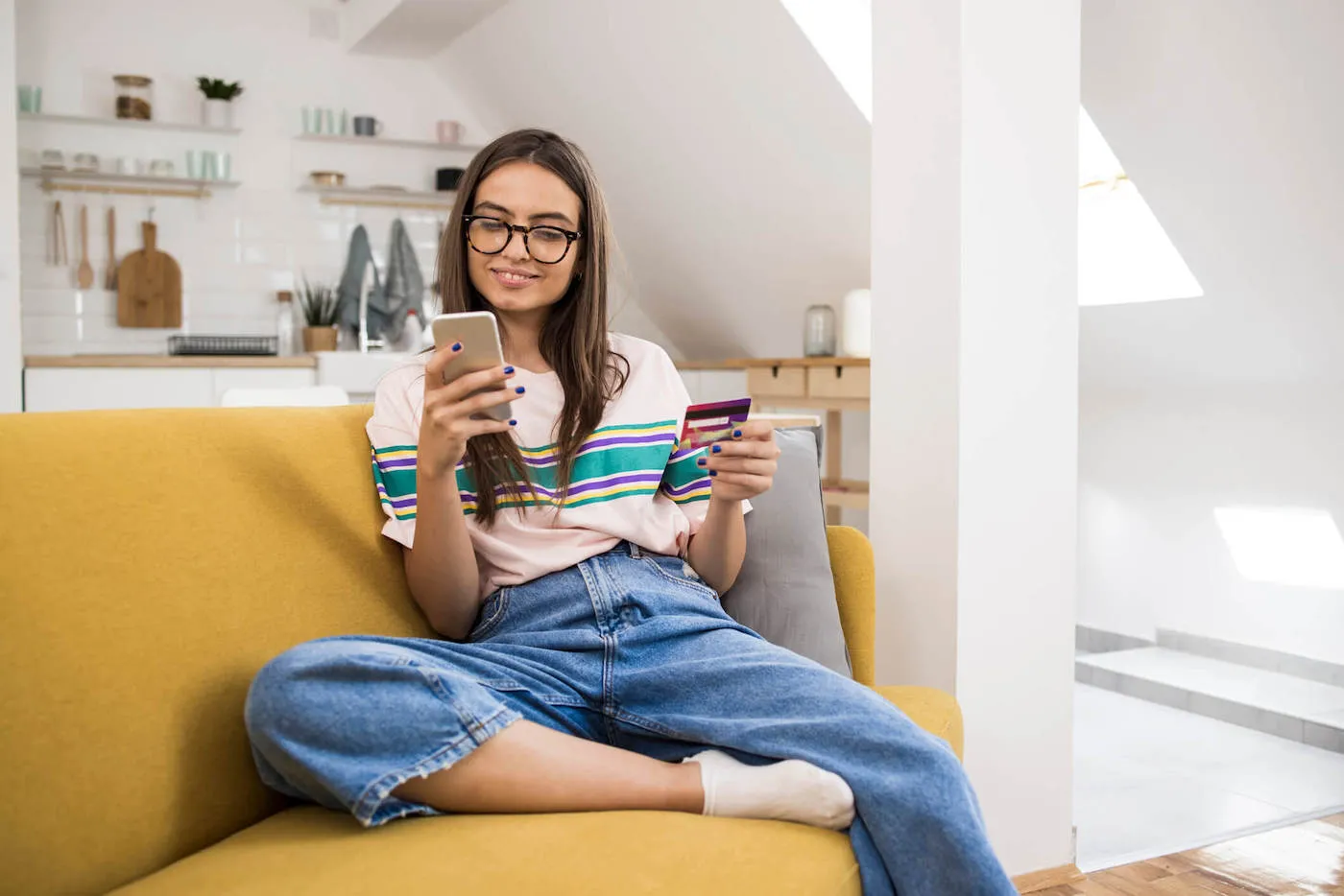 A young women sits on her yellow couch as she smiles at her phone while holding her credit card.