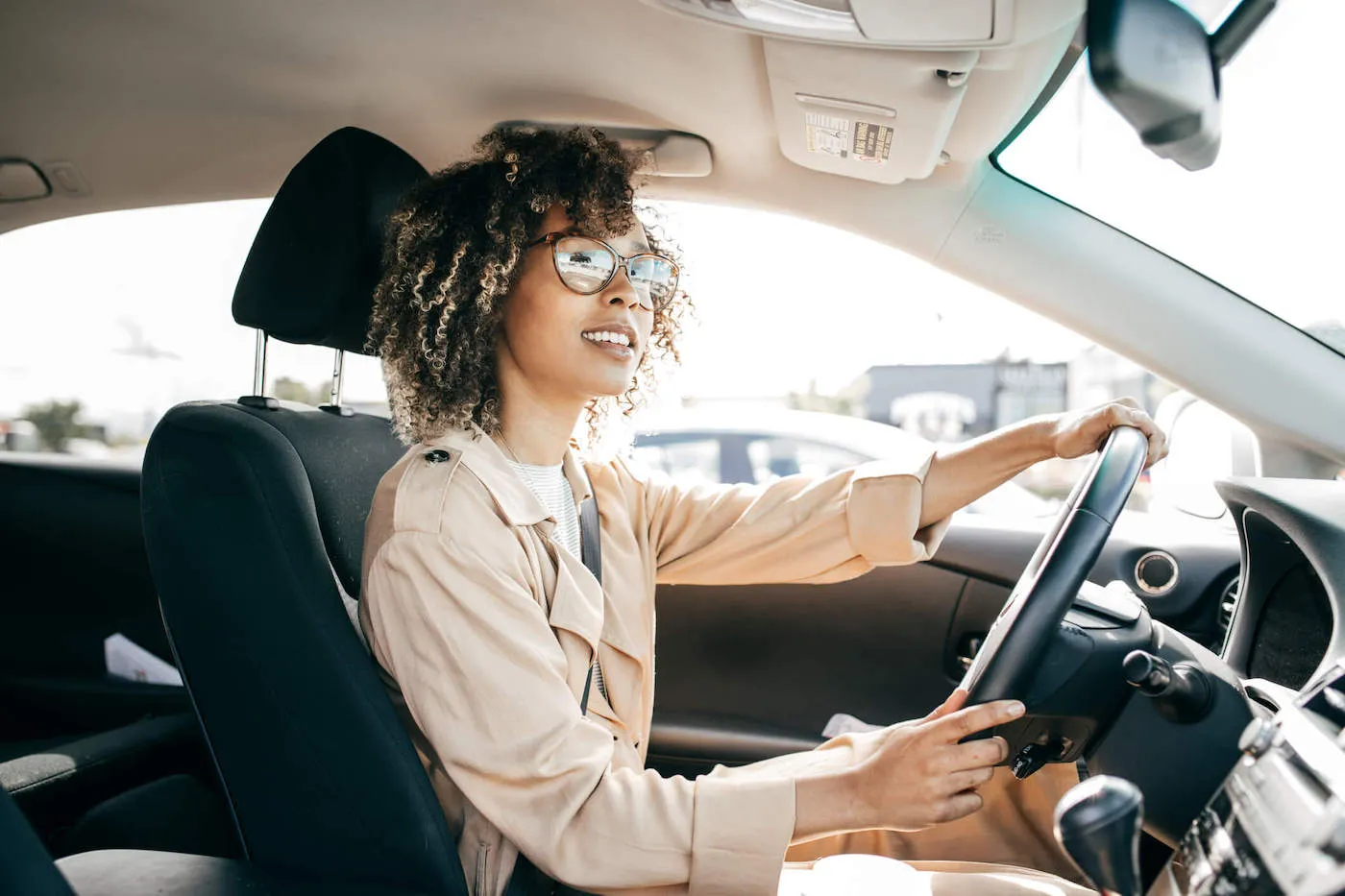 A young women with brown glasses and curly hair is driving her car while wearing a beige coat.