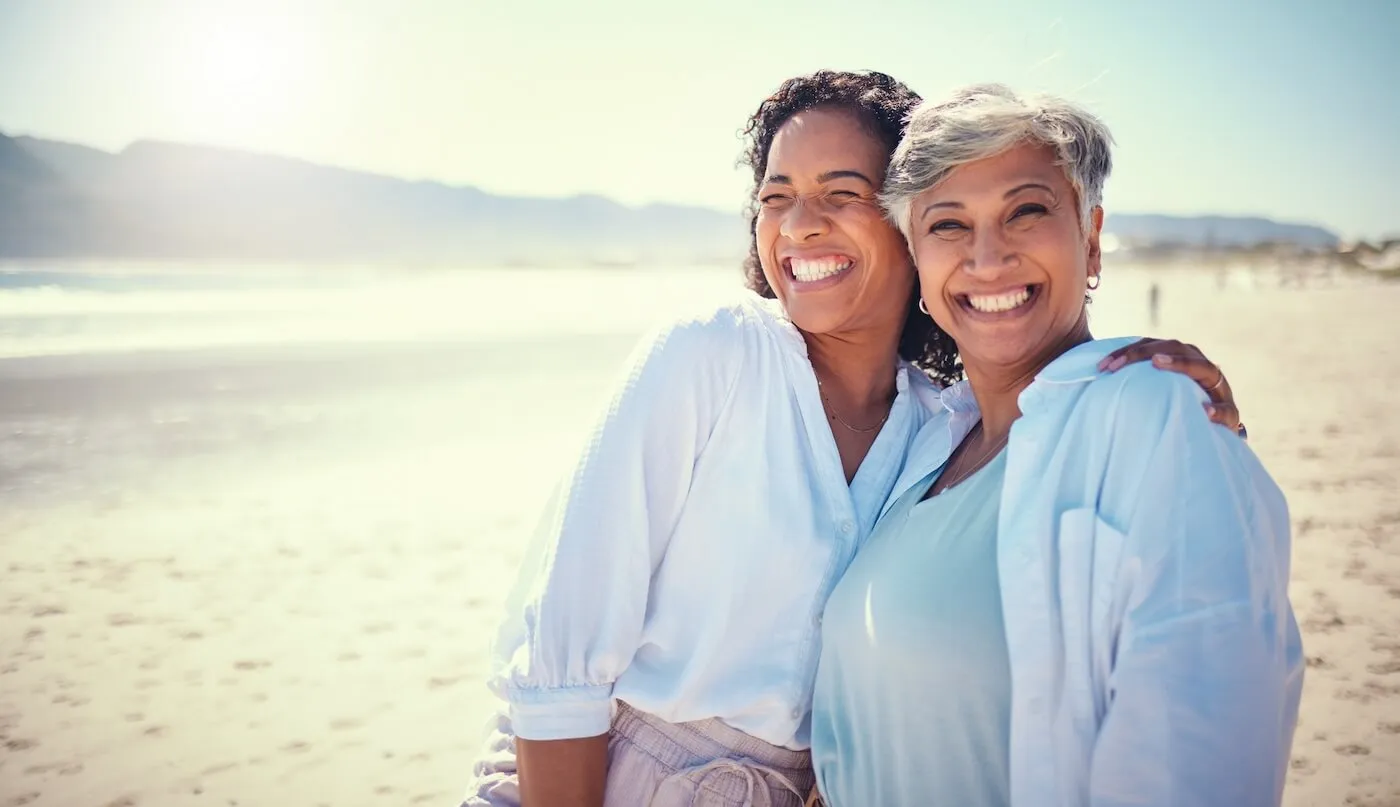 A mother and adult daughter smiling while posing for a photo on a sunny beach