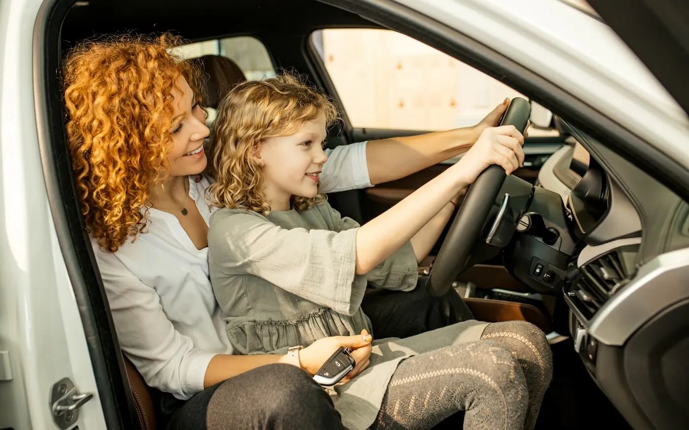 A woman is letting her daughter steer the wheel of a parked car