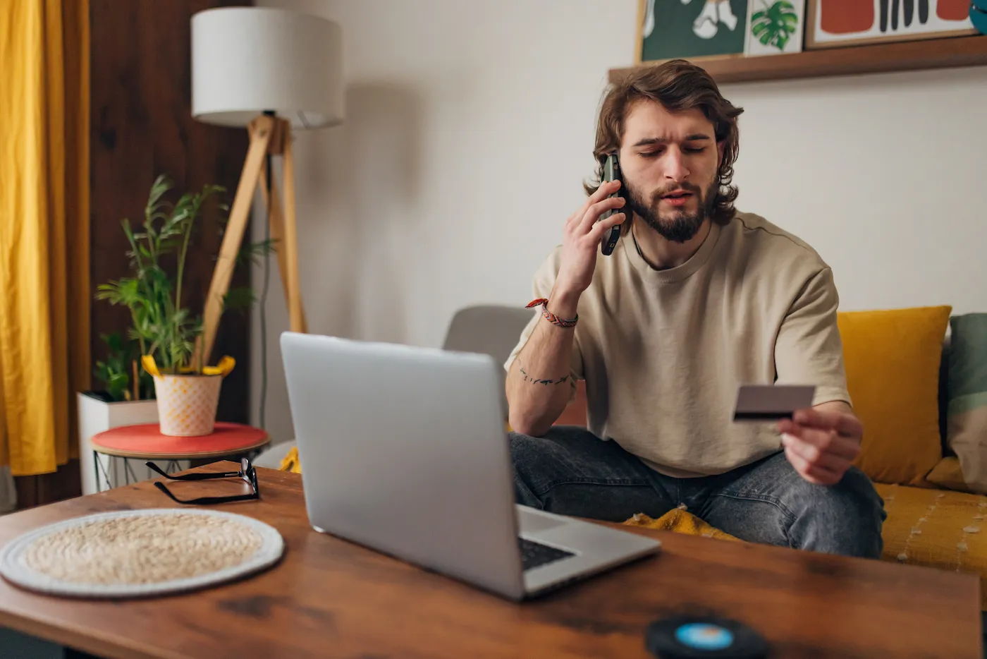 Man is sitting on a sofa in the living room, holding a credit card in his hands and making a phone call