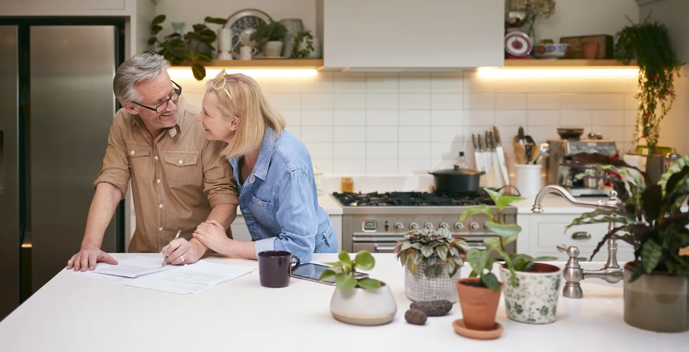 Mature Couple Reviewing And Signing Domestic Finances Paperwork In Kitchen At Home.