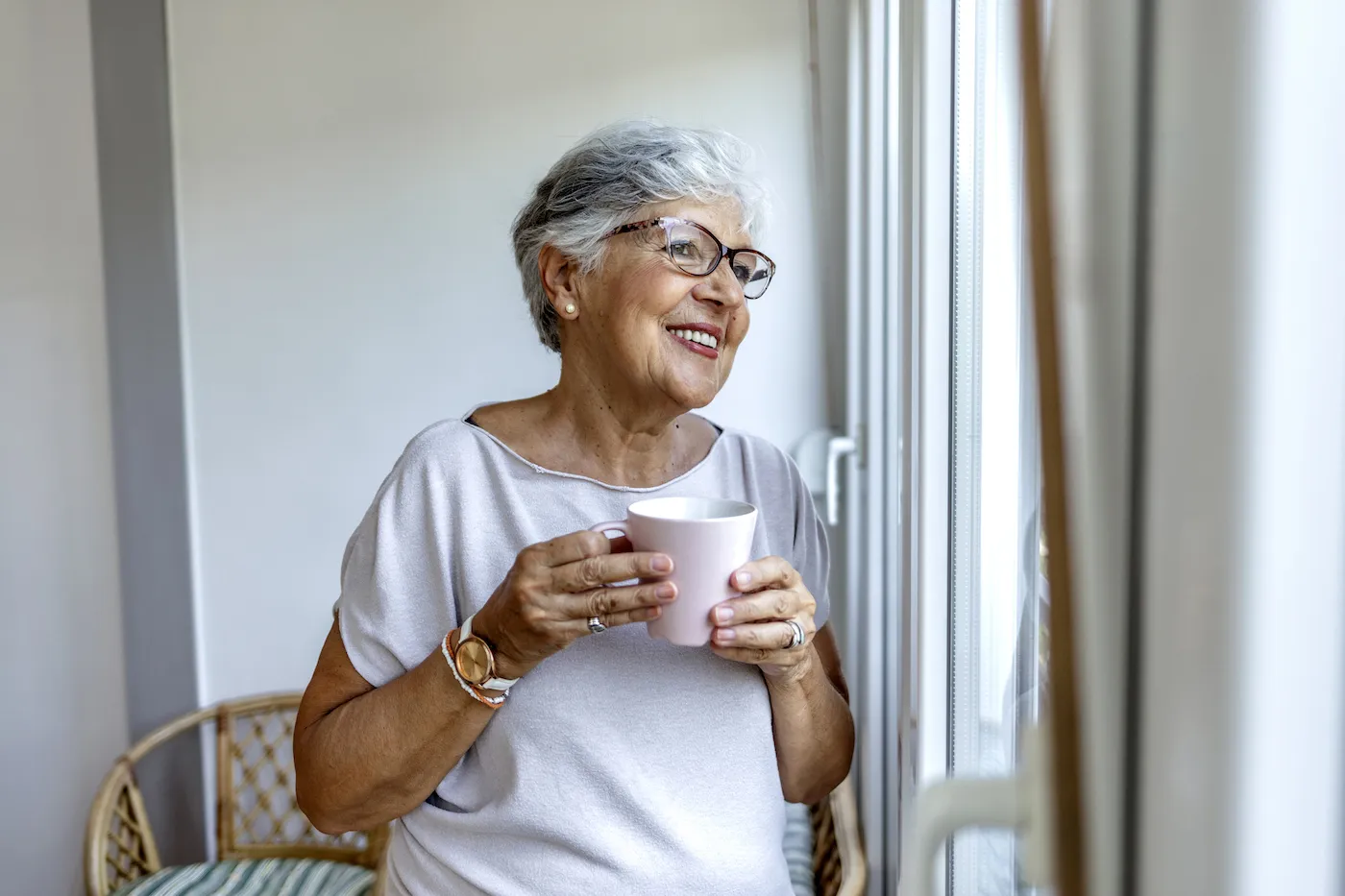 Mature woman standing happily in her home by a window enjoying some quiet time with a cup of tea.