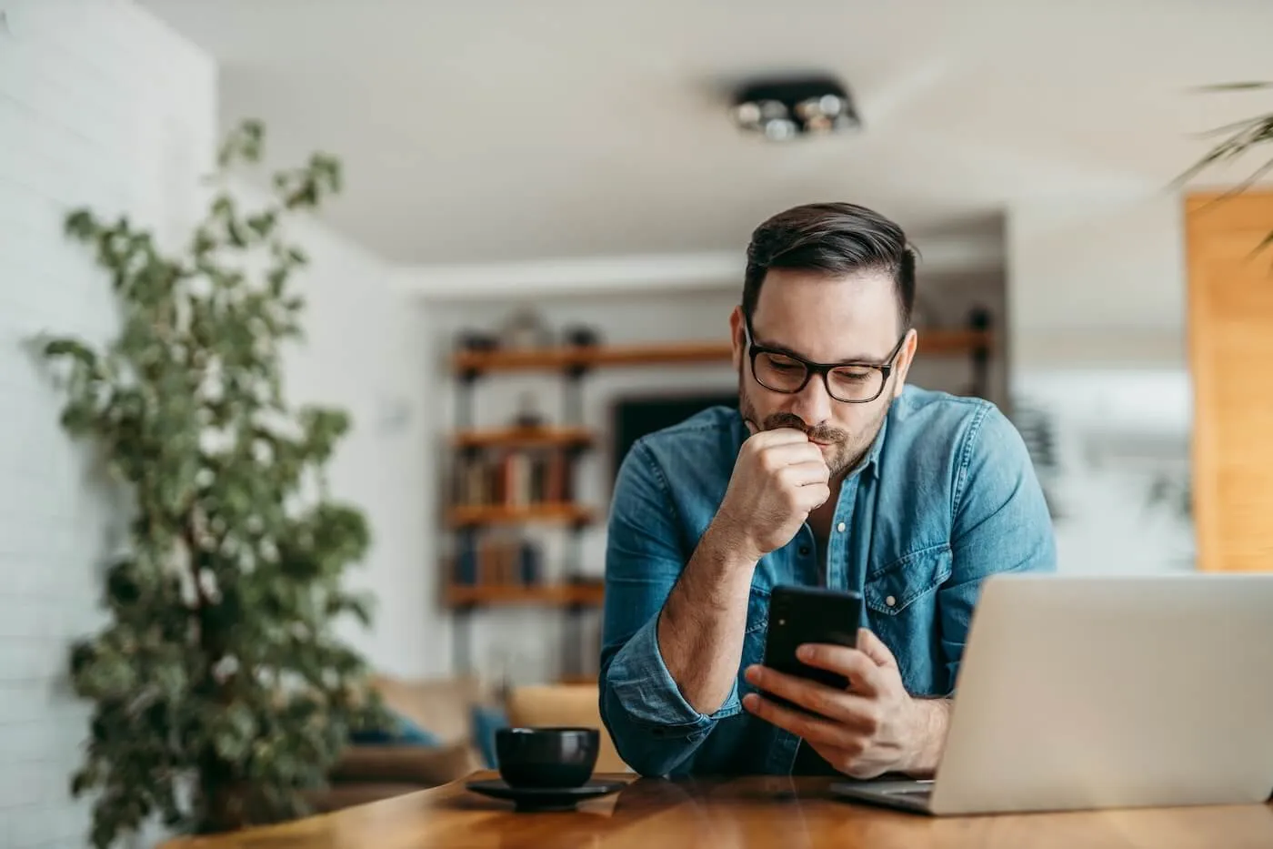 Focused man using his smartphone, with a laptop and a cup of hot drink on his desk