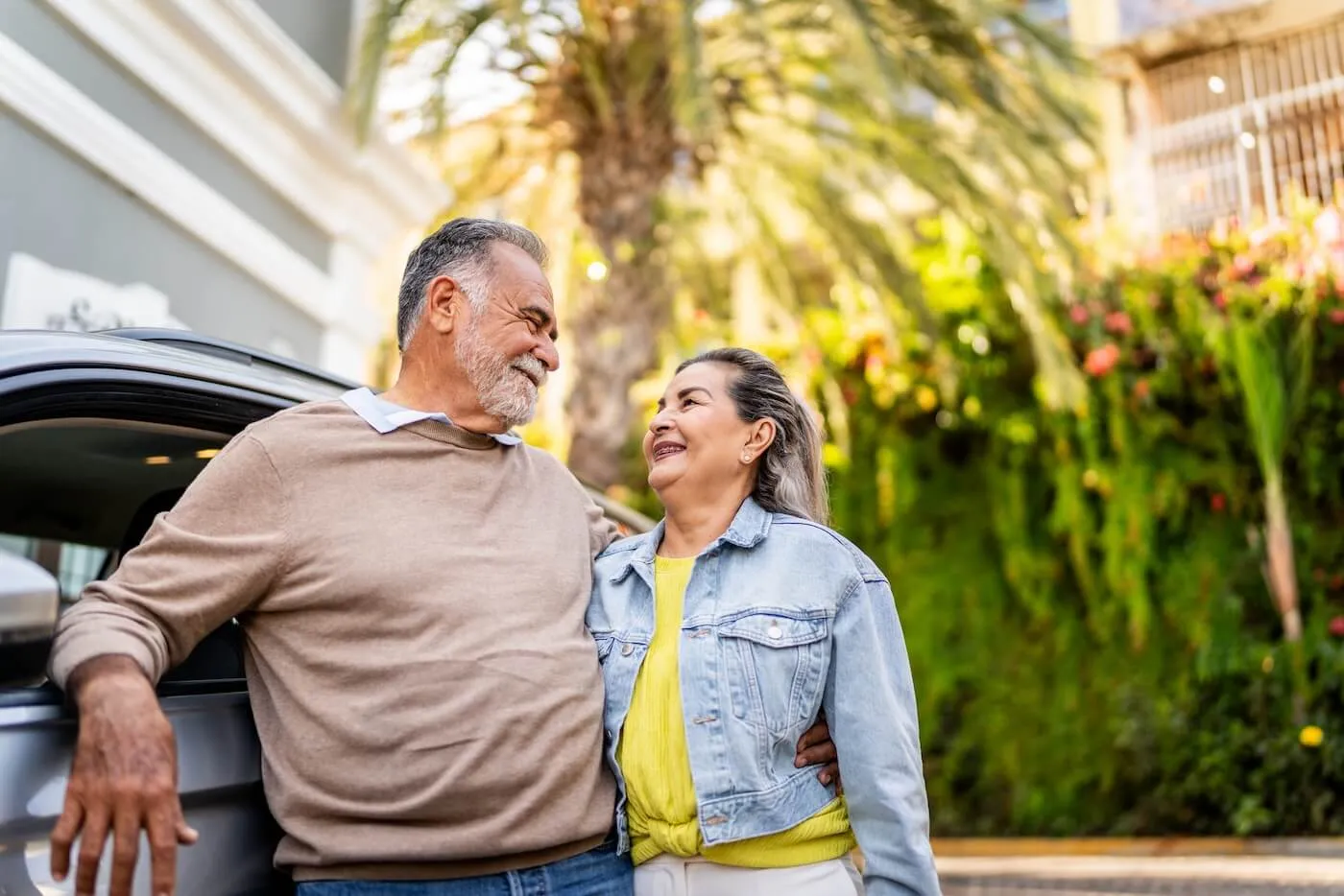 Smiling senior couple standing next to their car and looking at each other. The tropical plants grow on the background.