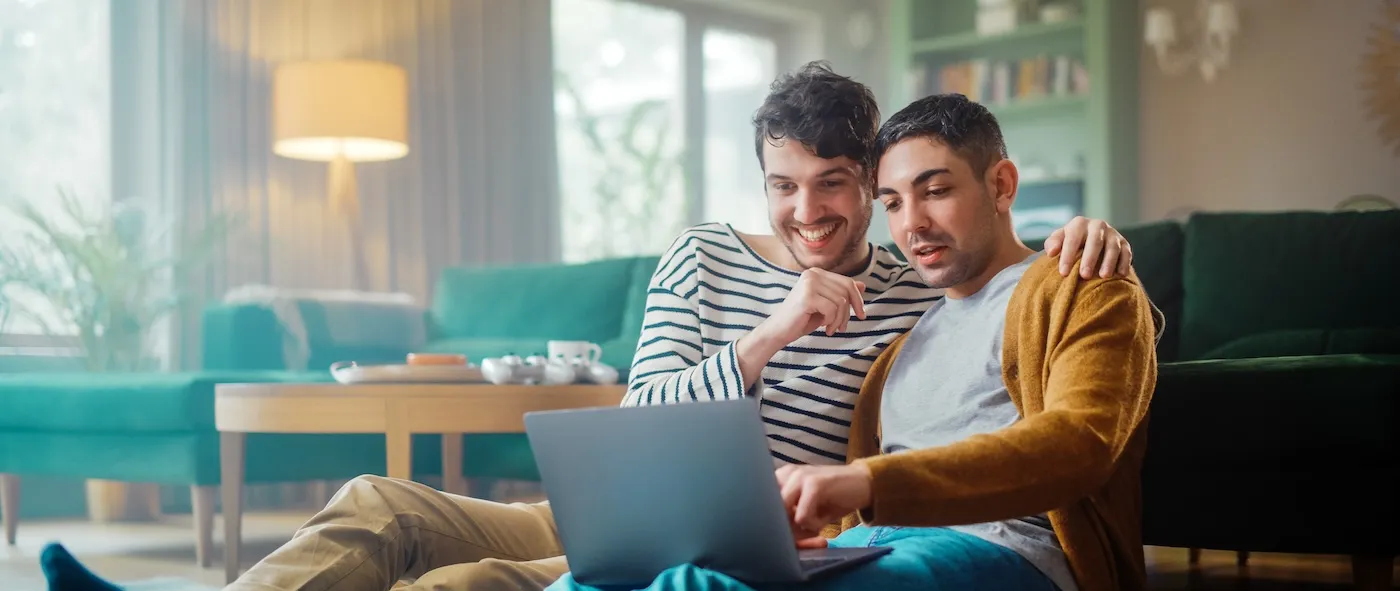 A Handsome Gay Couple Using Laptop Computer, while Sitting on a Living Room Floor in Cozy Stylish Apartment.