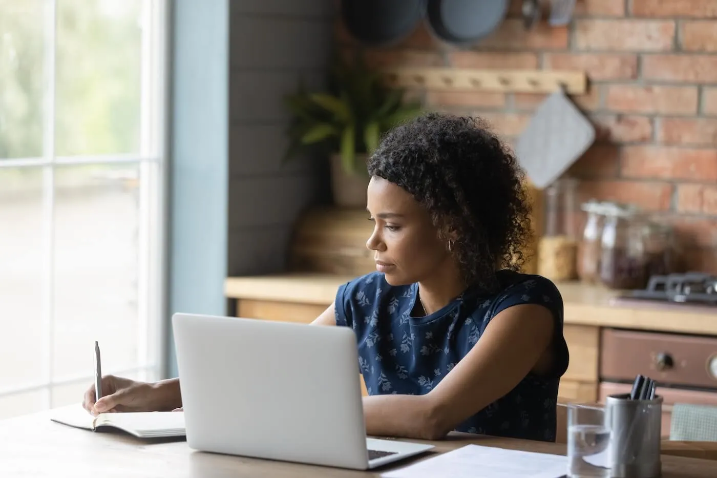 Focused woman making notes while using her laptop in the kitchen