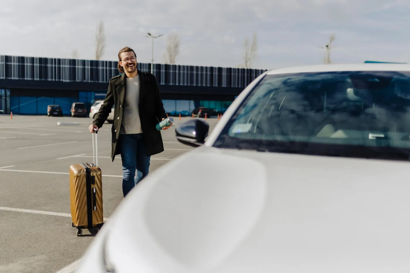 Smiling man with a suitcase and a water bottle approaching the rental car next to the airport