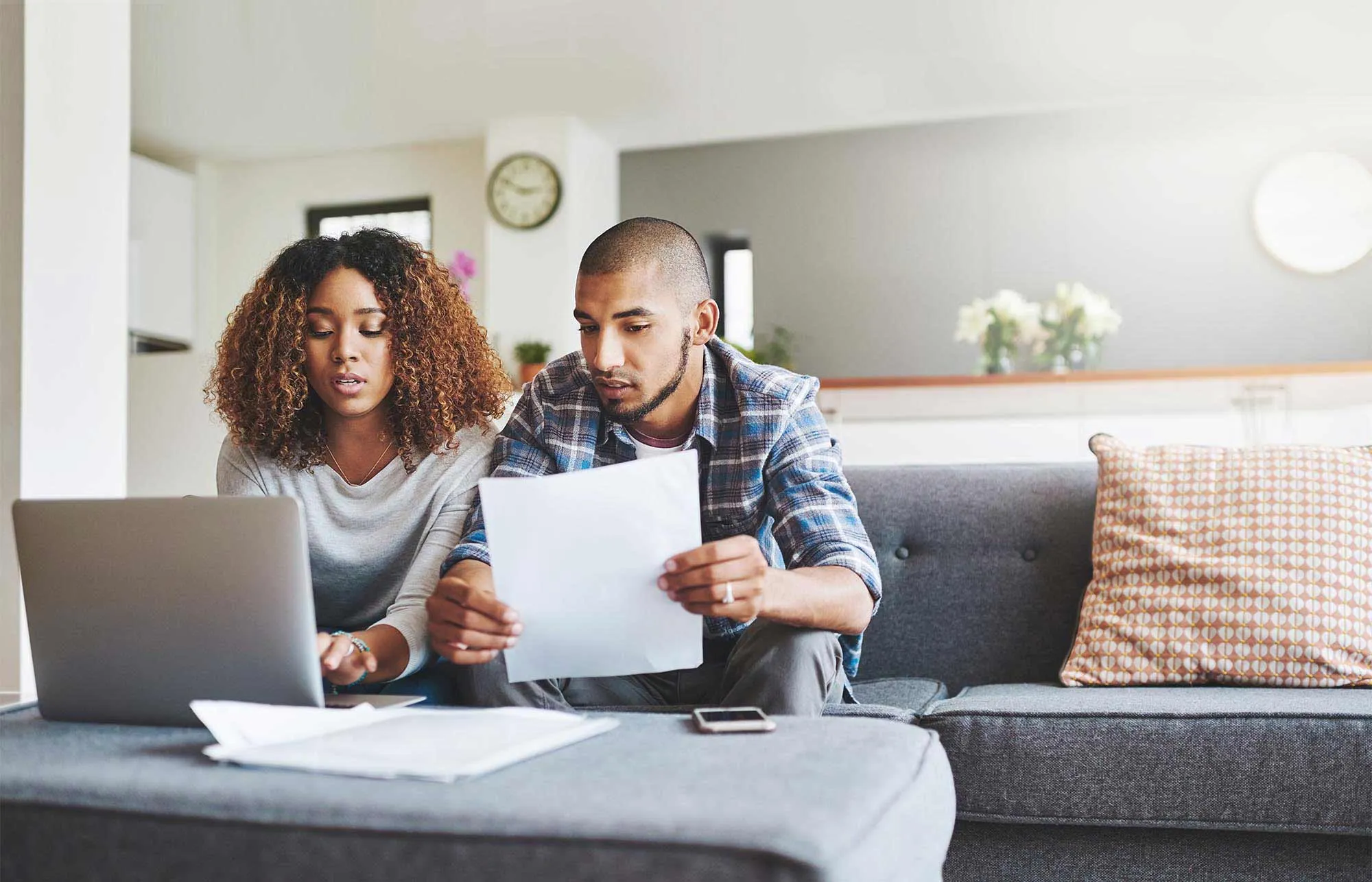 A couple sits on the couch while looking at a laptop computer while the man holds a document in his hands.