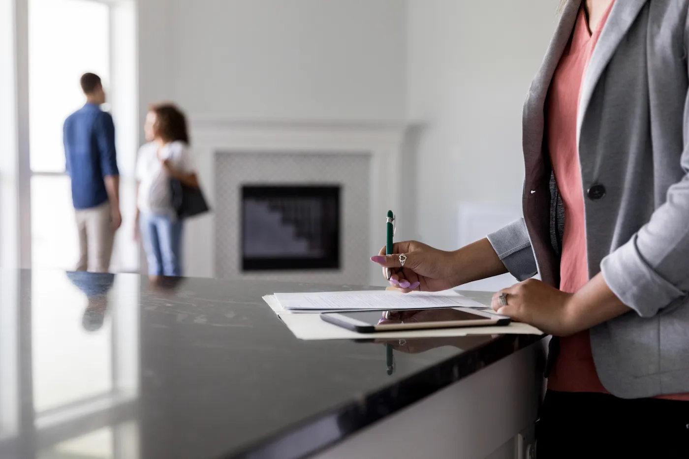 Female real estate agent finishes up paperwork as a couple discuss the purchase of a new home in the background.