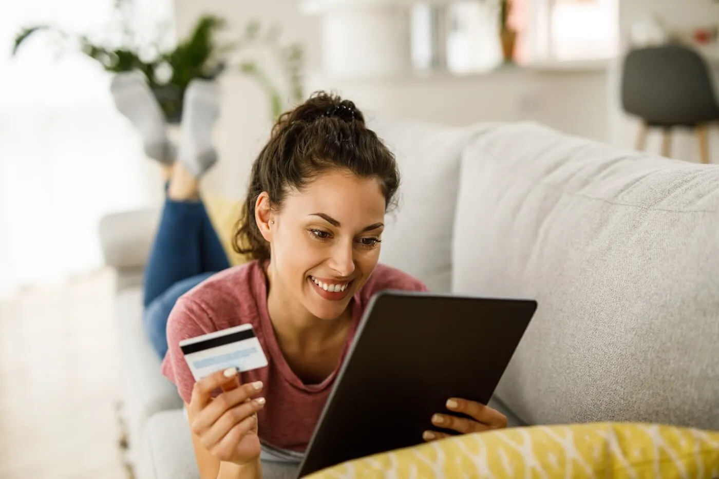 Smiling woman lying on a couch and holding a credit card while checking her score on a tablet