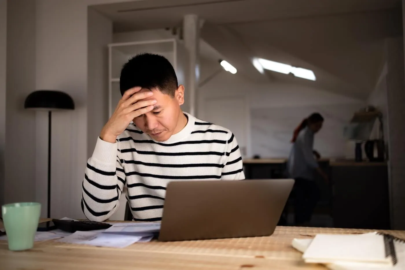 Concerned man reviewing the bills while using the laptop, his wife is on the kitchen on the backgroung