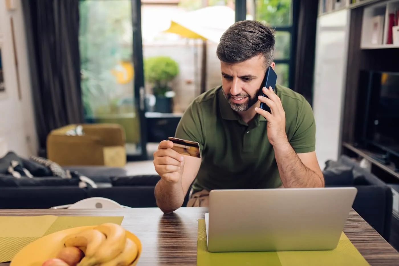 Man is making a phone call while looking at his credit card and using a laptop