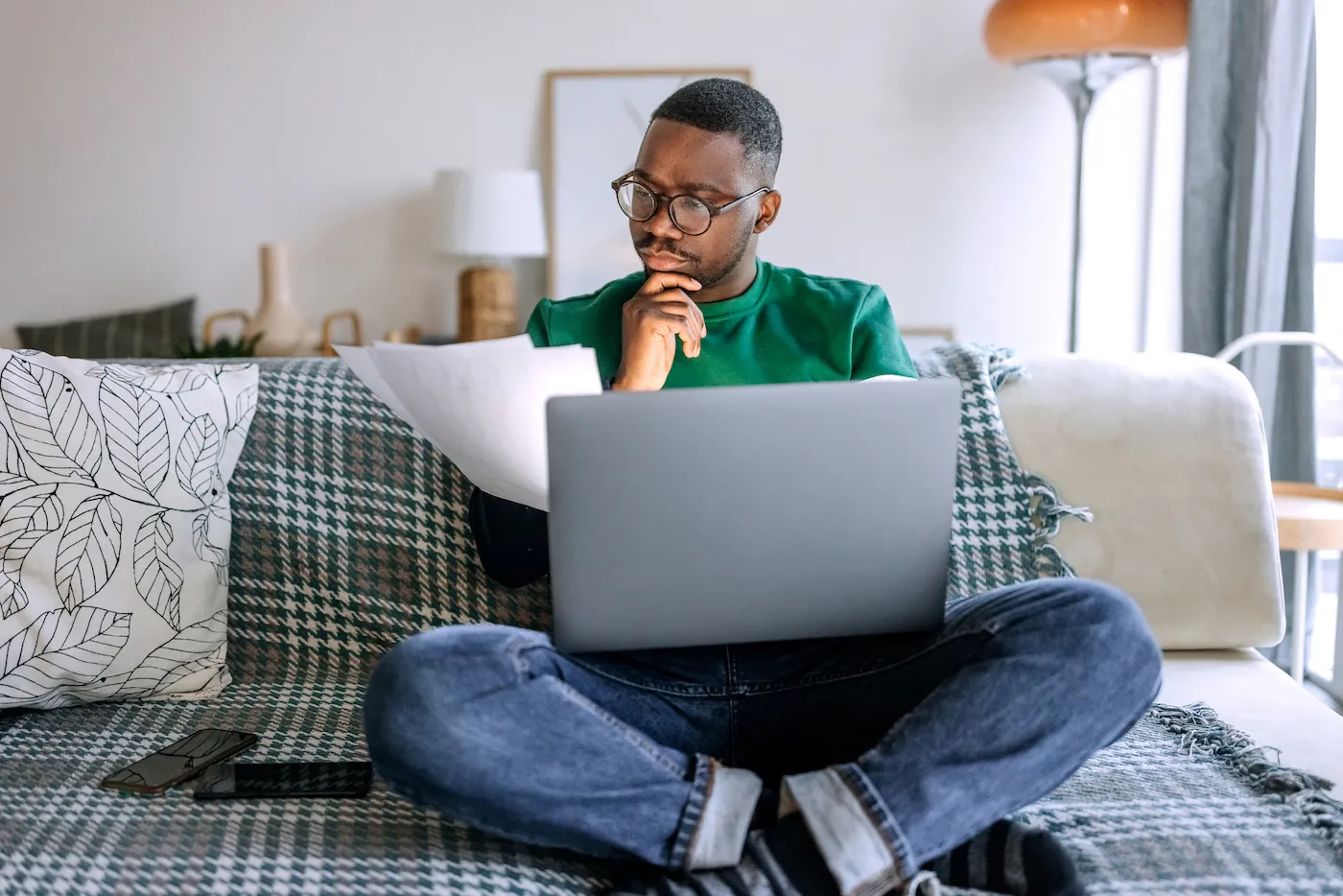 Young concentrated man going over paperwork while working from living room, he is sitting on the sofa with laptop in his lap.