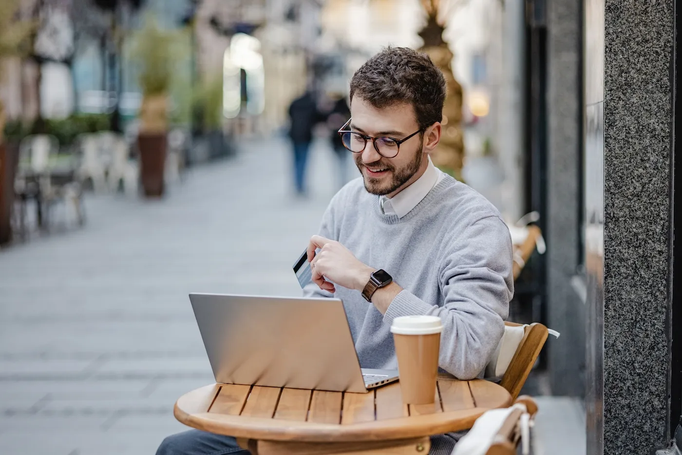 A young man using a laptop and holding a credit card while sitting at an outdoor cafe.