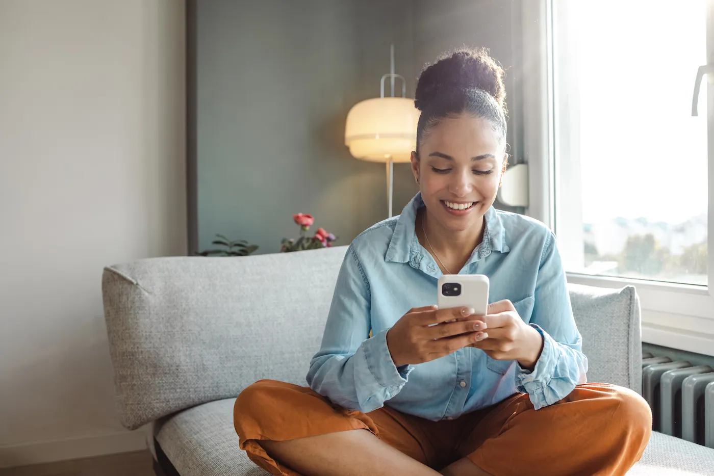 A young woman is at home in the living room, she is using a mobile phone and smiling