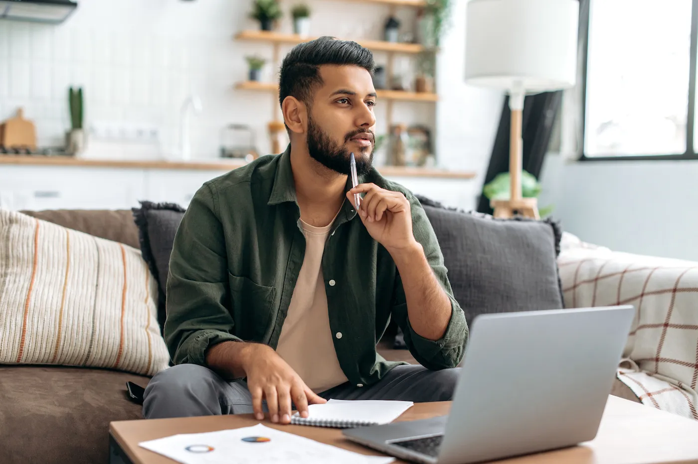 Pensive guy, using laptop, sits at home on sofa, looks away, thinking