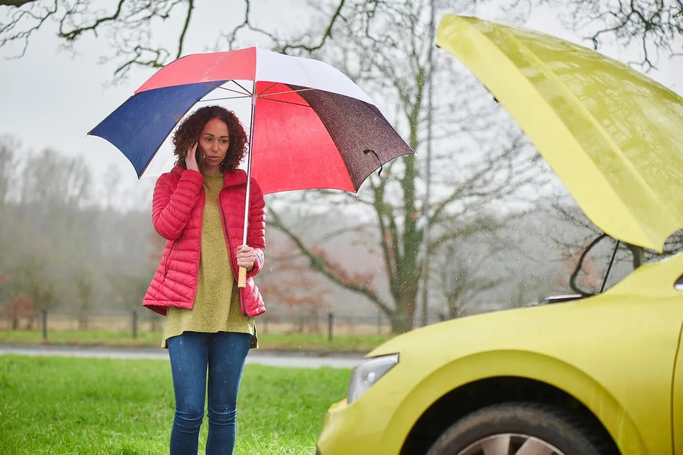 Concerned woman with umbrella is making a phone call while looking at the car with an open hood