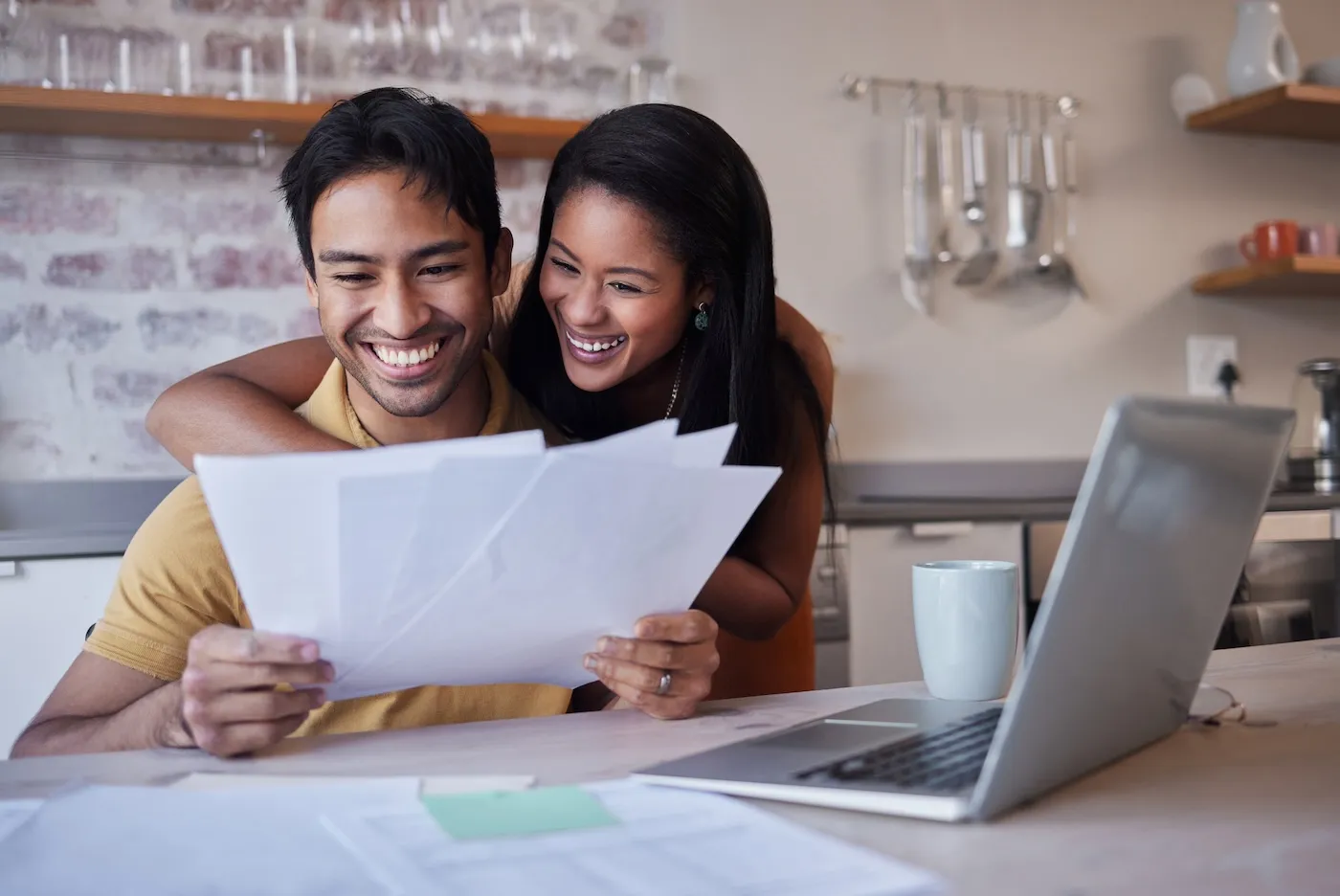 Happy married couple reviewing car insurance documents at the kitchen table in front of a laptop.