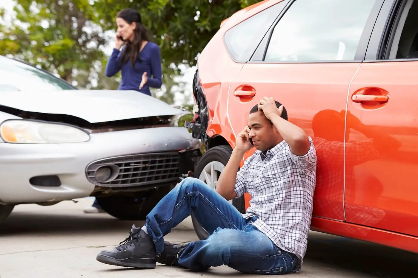 Frustrated man is sitting on the ground and leaning against a red car while calling the insurance company after the collision. A female driver of a silver car is also making a phone call.