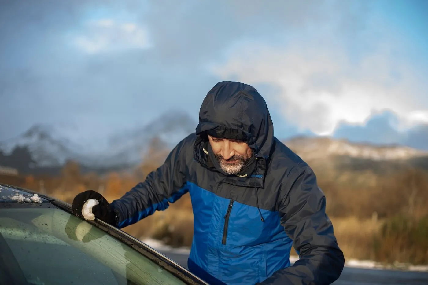 Concerned mature man in a winter jacket assessing the hail damage of his car in a mountain area