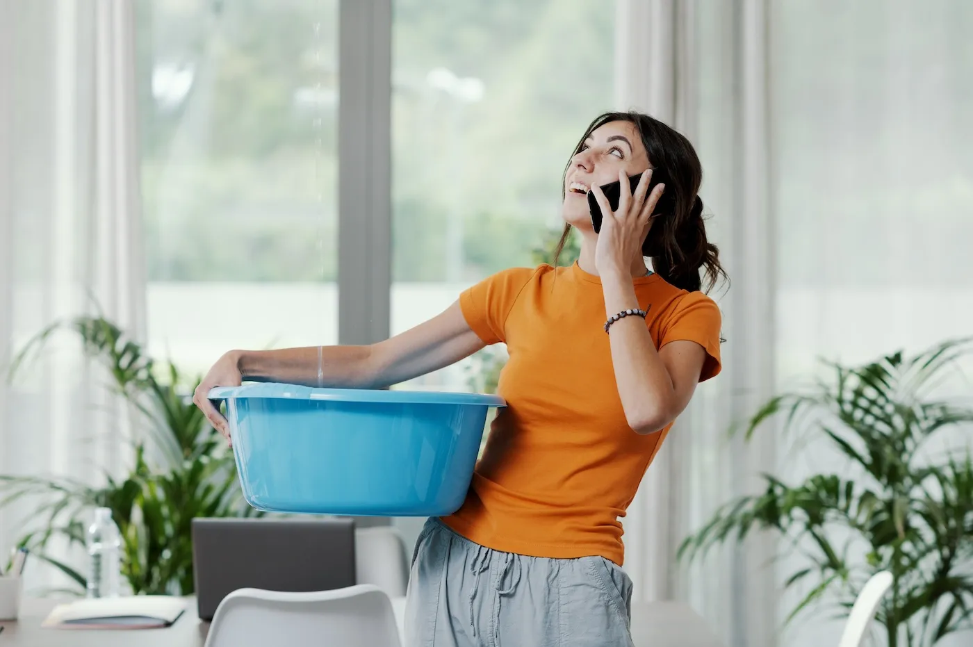 Young upset woman collecting water leaking and dripping from the ceiling as she talks on the phone.