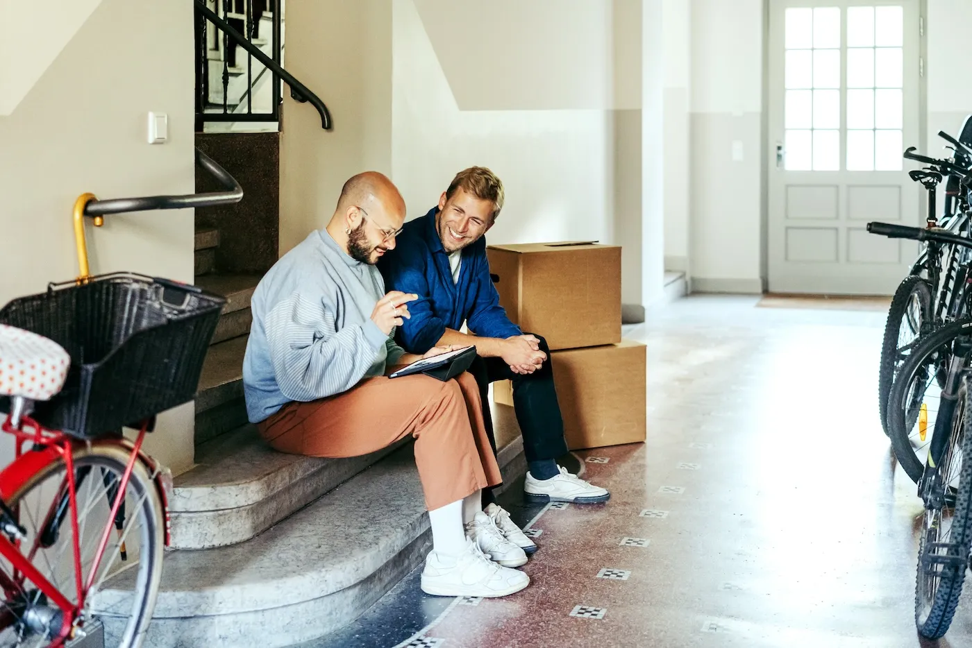 Two young men sitting on step with cardboard boxes using digital tablet for choosing moving service for house relocation.