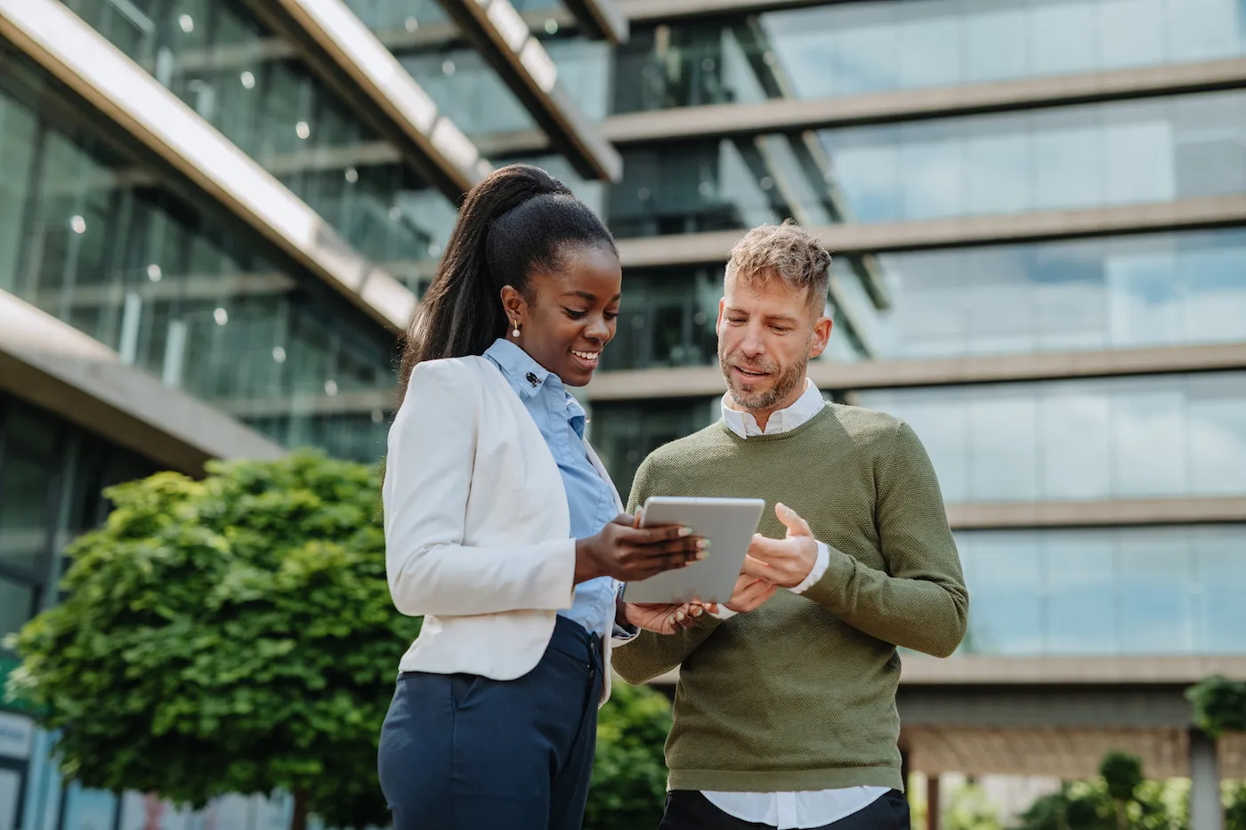 Two business people in financial district looking at investing documents together. They are standing outside a large office building with trees.