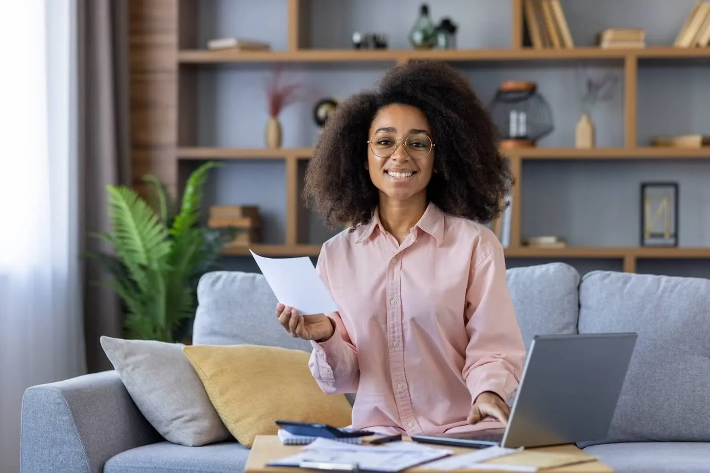 Smiling woman managing finances at home, using a laptop for budgeting and holding a document.